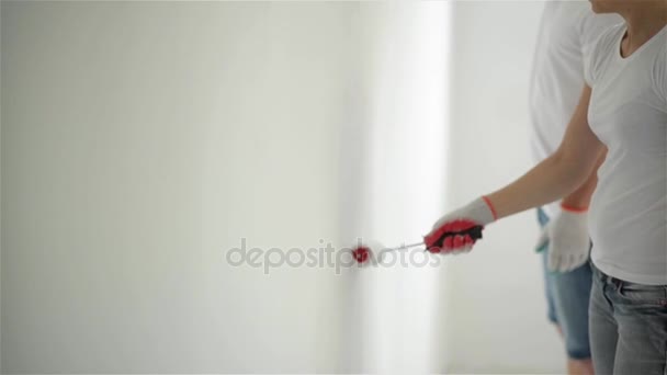 Interni Ritratto di coppia felice attiva che fa la riparazione dell'appartamento insieme. Giovane ragazza e uomo in camicie e jeans dipingere il muro con vernice bianca . — Video Stock