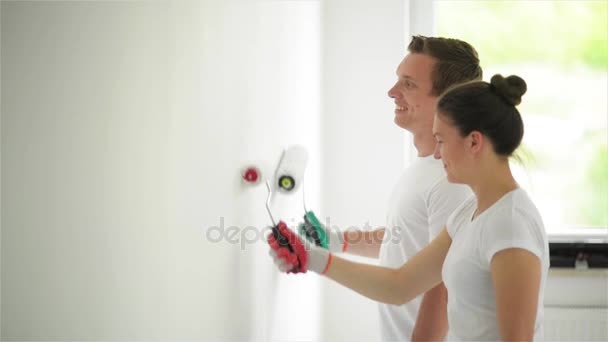 Вид сбоку счастливой улыбающейся пары, рисующей стену белой краской в гостиной вместе. Женщина и мужчина носят яркие рубашки и держат платья в руках . — стоковое видео