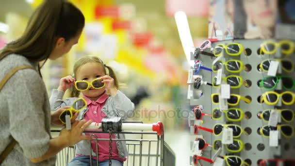 Interior retrato de feliz fêmea criança com ela bonita mãe escolher óculos de sol no supermercado . — Vídeo de Stock