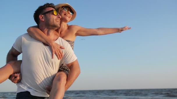 Lachen gelukkig paar genieten van mooi weer in de buurt van de zee. Sterke Man met zijn vriendin op de schouders op de blauwe hemelachtergrond. — Stockvideo