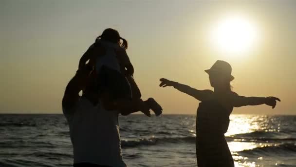 Dunkle Silhouetten von Mutter mit Sommerhut und Vater mit kleiner Tochter am Meer bei Sonnenuntergang. starker Mann wirbelt Mädchen über den Kopf. — Stockvideo