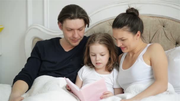 Pretty Little Girl está tratando de leer con sus padres acostados en la cama. Hermosa niña está leyendo mostrando por dedo algo en el libro . — Vídeos de Stock