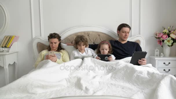 Los padres y los niños usan dispositivos móviles juntos en el dormitorio durante el fin de semana en casa. Pareja joven con niños están acostados en la cama sosteniendo en las manos aparatos electrónicos y hablando entre sí . — Vídeo de stock