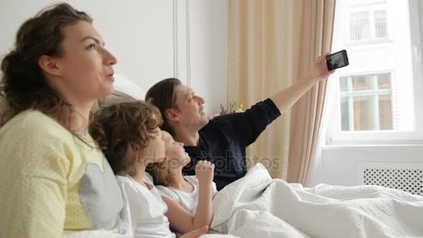 Homem bonito está tirando uma foto por seu smartphone. Selfie família positiva com mãe, pai e duas crianças deitadas na cama e vestindo pijamas . — Vídeo de Stock