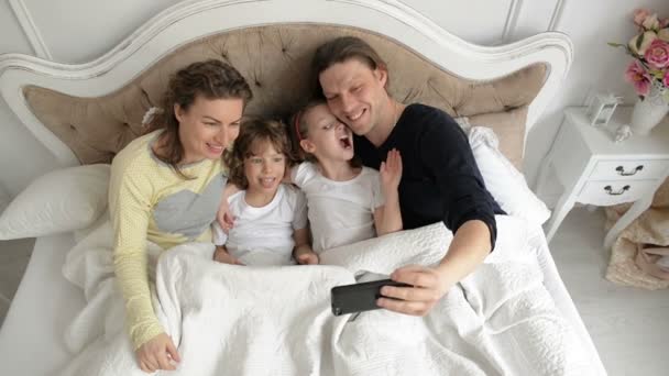 Joyous Family is Taking Selfie Using Smartphone. Cute Boy and Girl are Kissing Their Parents Lying on the Bed. — Stock Video