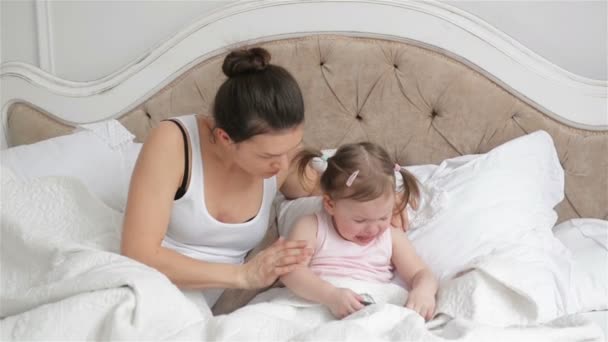 Mutter versucht ihre Tochter im Schlafzimmer zu beruhigen und reicht ihr Smartphone. niedliches kleines Mädchen mit zwei Pferdeschwänzen weint auf dem Bett sitzend. — Stockvideo