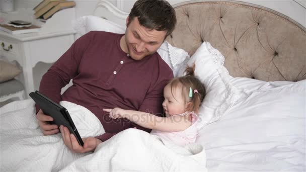 La Familia Moderna se Relaja en el Fin de Semana del Dormitorio Juntos. Padre joven e hija pequeña están usando aparatos acostados en la cama . — Vídeos de Stock