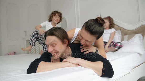 Os pais jovens com sorrisos encantadores estão gastando tempo livre com seu filho e filha no quarto na manhã. Família inquieta e ativa brincando juntos em uma cama em casa durante Weeked . — Vídeo de Stock