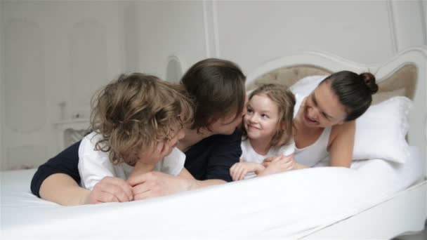 Pareja joven con niños divirtiéndose juntos en el dormitorio. Padres juguetones están disfrutando de fin de semana con su hijo y su hija acostados en la cama blanca . — Vídeos de Stock