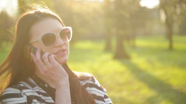 Портрет красивой женщины со смартфоном на открытом воздухе. Улыбающаяся брюнетка в длинных волосах и модных солнцезащитных очках разговаривает по мобильному телефону во время солнечного весеннего дня в парке . — стоковое видео