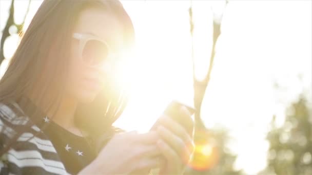 Porträt einer lächelnden Brünetten mit Smartphone im Park. niedliche Frau mit Sonnenbrille und langen Haaren nutzt mobiles Gerät und genießt Sonnenstrahlen und warmen Tag im Freien. — Stockvideo
