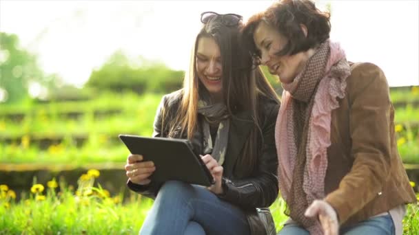 Retrato de mujeres felices con dientes blancos riendo y sosteniendo la tableta en las manos. Lindas morenas se están divirtiendo junto con dispositivo electrónico en el parque . — Vídeo de stock