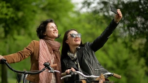Insouciantes Les femmes aux cheveux foncés prennent des photos d'elles-mêmes à l'aide d'un téléphone intelligent à l'extérieur. Deux jolies filles avec des vélos faisant Selfie par téléphone dans le parc . — Video