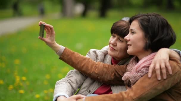 Szczęśliwa rodzina na piknik w parku przy użyciu telefonu komórkowego do podjęcia Phote siebie. Dorosły córka z jej matka są biorąc Selfie przez smartfon na zewnątrz. — Wideo stockowe