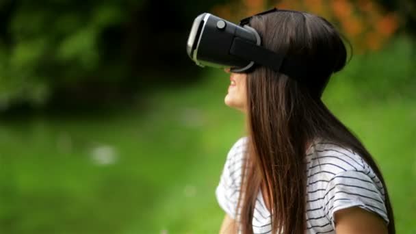 Primer plano Retrato de la joven mujer bonita con camisa a rayas está utilizando gafas de realidad virtual sentado cerca del lago en el parque de la ciudad . — Vídeos de Stock
