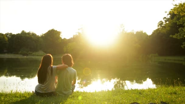 Pohled zezadu na romantický mladý pár sedí na zelené trávě poblíž krásné jezero a těší Sunset. — Stock video