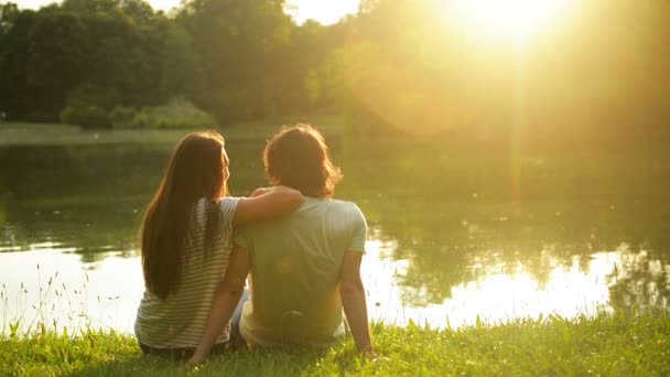 Hombre guapo y mujer guapa mirando el agua del lago y la puesta del sol. Vista trasera de una pareja encantadora romántica en el exterior . — Vídeos de Stock