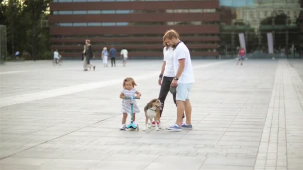 Ευτυχισμένη οικογένεια το περπάτημα σε όλη την πλατεία της πόλης με ένα σκυλί. Μικρό κορίτσι στο φόρεμα βόλτες σε ένα σκούτερ. Ο πατέρας πηγαίνει με ένα λαγωνικό. — Αρχείο Βίντεο