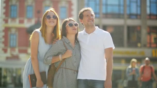 Drie lachende vrienden uitgaven tijd samen buiten tijdens een warme zomerdag. Man dragen witte Shirt staat op het oude stadsplein met twee meiden in modieuze zonnebrillen en jurken. — Stockvideo