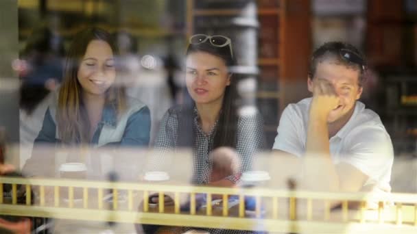 Grupo de jovens se divertindo em um café e esperando por algumas bebidas. Duas mulheres e um homem no restaurante visto através da janela na rua . — Vídeo de Stock