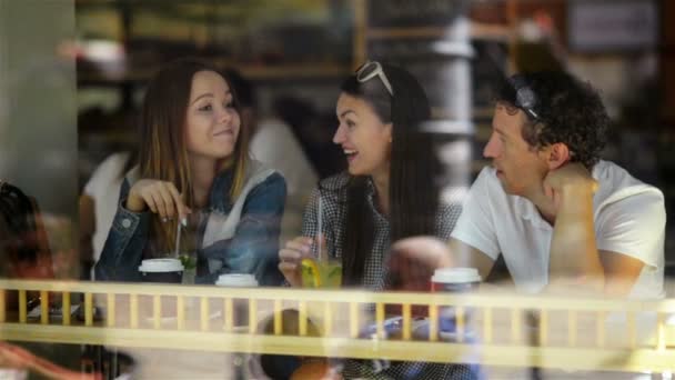 Три счастливых друга пьют и хорошо проводят время за обеденным столом в кафе с коктейлями. Один мужчина и две красивые женщины сидят у большого окна в ресторане . — стоковое видео