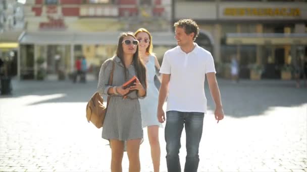 Tre attraktiva ungdomar möttes i gatan och de är glada att tillbringa tid tillsammans. Två flickor i solglasögon och klänningar och en pojke i vit skjorta leende och omfamna utomhus. — Stockvideo