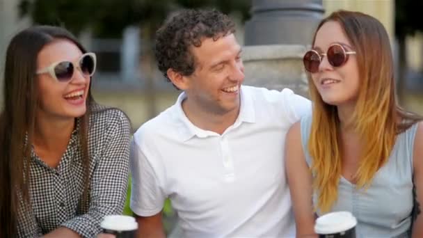 Primer plano Retrato de tres amigos sonrientes sentados con tazas de café al aire libre y hablando durante el verano. Dos chicas con un chico bebiendo bebidas afuera . — Vídeo de stock