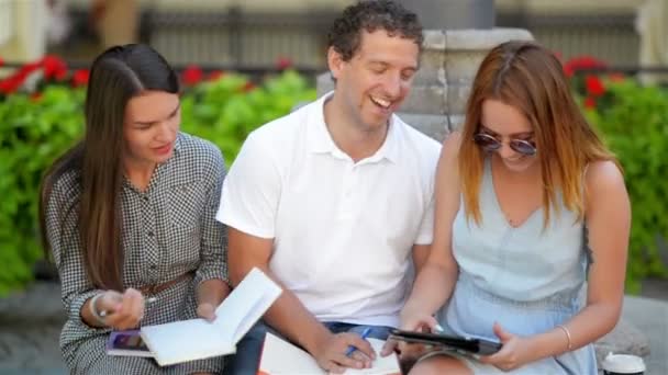 Två kvinnliga studenter med en pojke studerar grupp utomhus sitter på bänken i stadsparken. E-learning koncept. — Stockvideo