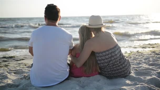 Madre, Padre e Figlia seduti sulla spiaggia vicino al mare e abbracciati guardando il sole e le onde . — Video Stock