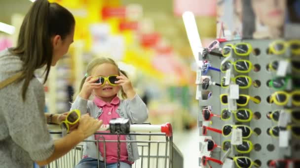 Menina Liitle bonito com dois rabos de cavalo sentado em um carrinho de compras e escolhendo óculos de sol no supermercado com sua mãe morena . — Vídeo de Stock