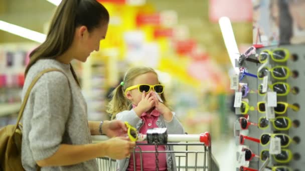 Szép kislány kiválasztása a napszemüveg a szupermarketben. Nő hosszú, sötét haj, és a sárga hátizsák segít neki kiválasztani. — Stock videók