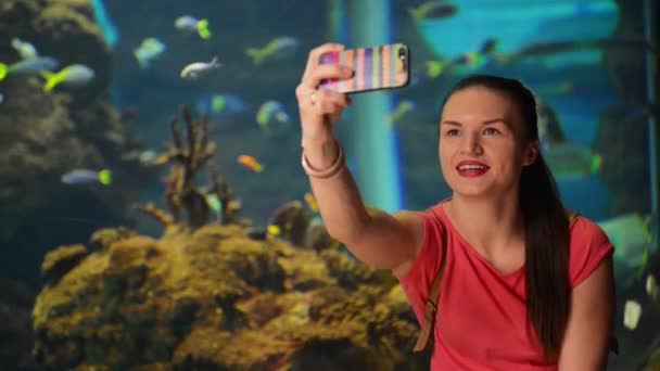 Junges Mädchen macht Selfie. Aquarium mit Fischen im Hintergrund. sie ist sehr glücklich und fröhlich und hat eine Menge Spaß. — Stockvideo