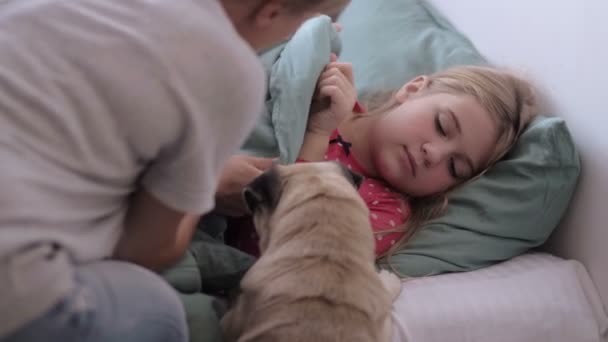 Мать ухаживает за больным ребенком лежа в постели с температурой гриппа с помощью термометра, девушка с лихорадкой лежа с милой собакой на диване дома, Семейное лечение — стоковое видео