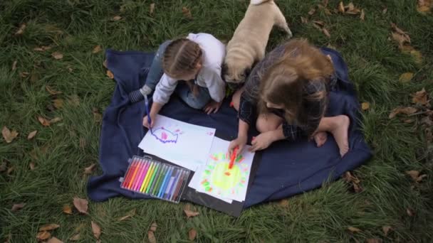 Zwei kleine Mädchen zeichnen in Notizbuch, während sie auf dem Gras im Park sitzen, Kinder zeichnen mit bunten Farben mit Mops Hund — Stockvideo