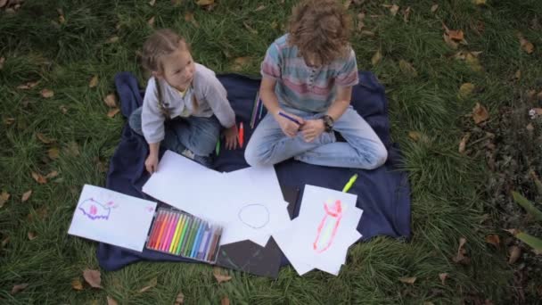 Két vidám gyerek, fiú és lány a zöld füvön fekszenek a nyári parkban. A gyerekek testvérei olvasnak, rajzolnak, játszanak, nevetnek.. — Stock videók