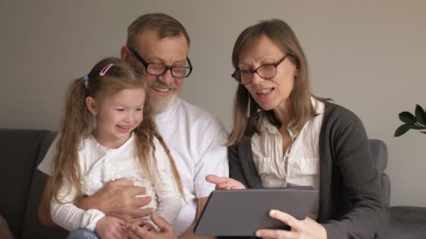 Concept de famille, de génération, de technologie et de personnes - grand-père, petite-fille et grand-mère souriants avec tablette assise sur le canapé à la maison — Video