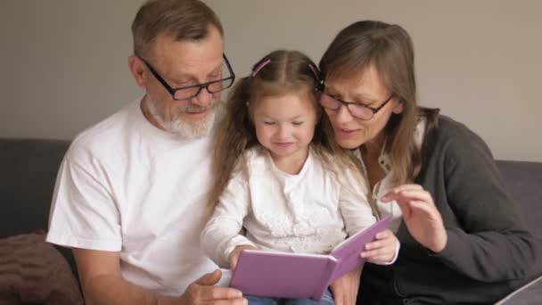 Familie, generatie en mensen concept - grootmoeder, grootvader en kleindochter lachende met boek zittend op Bank thuis — Stockvideo