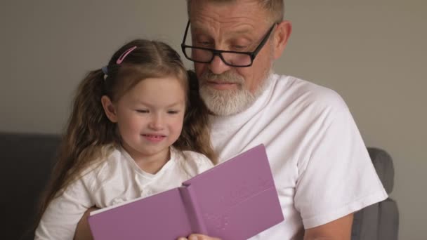 Портрет дедушки в очках и милая внучка-подросток, читающая книгу вместе, сидя дома на диване — стоковое видео