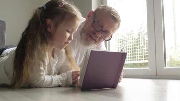 Leende tonåring flicka med senior man i glasögon läser bok tillsammans på golvet — Stockvideo