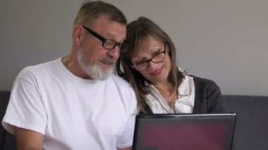Yaşlı bir çift gülüyor, uzaktan konuşuyor dizüstü bilgisayara bakıyor kanepeye oturmuş web kamerasına bakıyor olgun bir aile evde internet sohbetinin keyfini çıkarıyor.