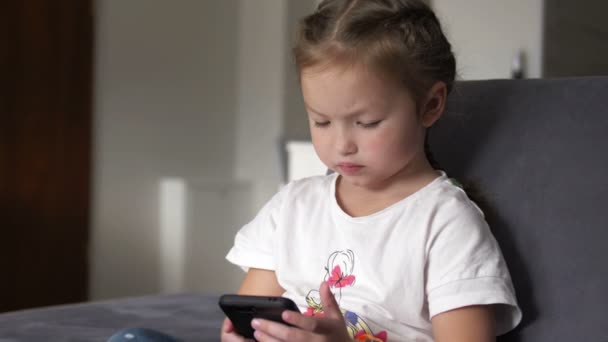 Criança casual sentada em um sofá em casa, brincando e tocando em um telefone celular — Vídeo de Stock