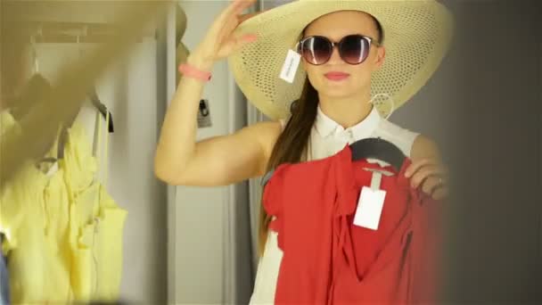 방 배경에 거울 근처 옷을 시도 하는 아름 다운 소녀입니다. 재미있는 모자와 선글라스를 착용한 젊은 여성. 해피 쇼핑 컨셉. — 비디오