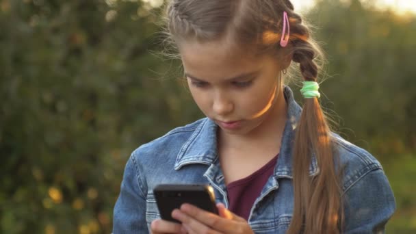 Kind gebruikt een mobiele telefoon. Het kind kijkt naar het scherm van de apparaten. Klein meisje speelt in kinderen digitale games in groen park of tuin buiten. — Stockvideo