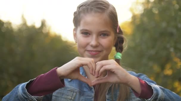 Menina adolescente bonita fazendo coração com as mãos ao ar livre. A olhar para a câmara. Infância . — Vídeo de Stock