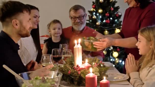 Glad familj firar semester tillsammans, sitter runt dekorerade runda bord. Mor som serverar middag. Vardagsrum med julgran, ljus och ljus — Stockvideo