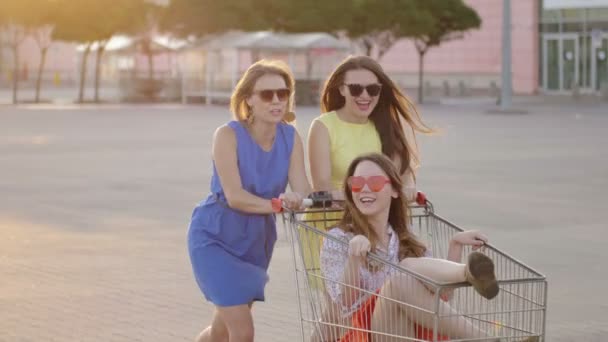 Freunde unterhalten sich bei hellem Tag auf dem Parkplatz des Einkaufszentrums. energische Frauen umkreisen attraktive fröhliche Mädchen im Einkaufswagen genießen Freizeit, slomo — Stockvideo