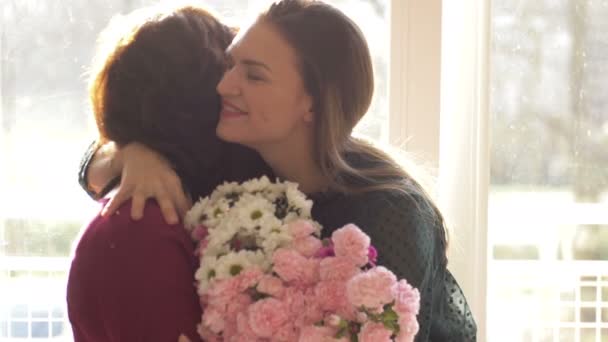 Молода доросла жінка дочка обіймає щасливу маму середнього віку, приймає зв'язок привітання зрілої мами з концепцією дня матері, представляє квіти, святкує разом — стокове відео