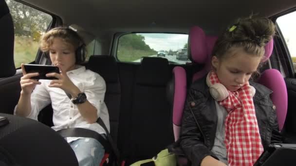Broder och syster använder telefonen och surfplattan när de reser i bilen. Pojke och flicka med hörlurar Spela en surfplatta och smartphone i auto under rida — Stockvideo