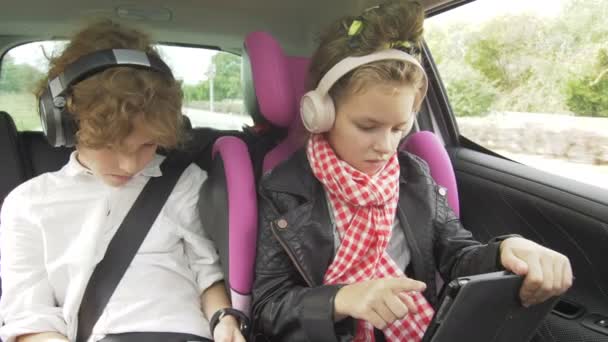 Menino e menina com fones de ouvido Jogando um tablet e smartphone em um carro, crianças usando um dispositivo no automóvel. Irmão e irmã viajando juntos — Vídeo de Stock