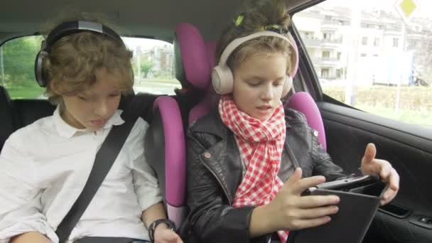 Хлопчик і дівчинка з навушниками Граючи планшет і смартфон в машині, діти використовують пристрої в авто. Брат і сестра подорожують разом — стокове відео